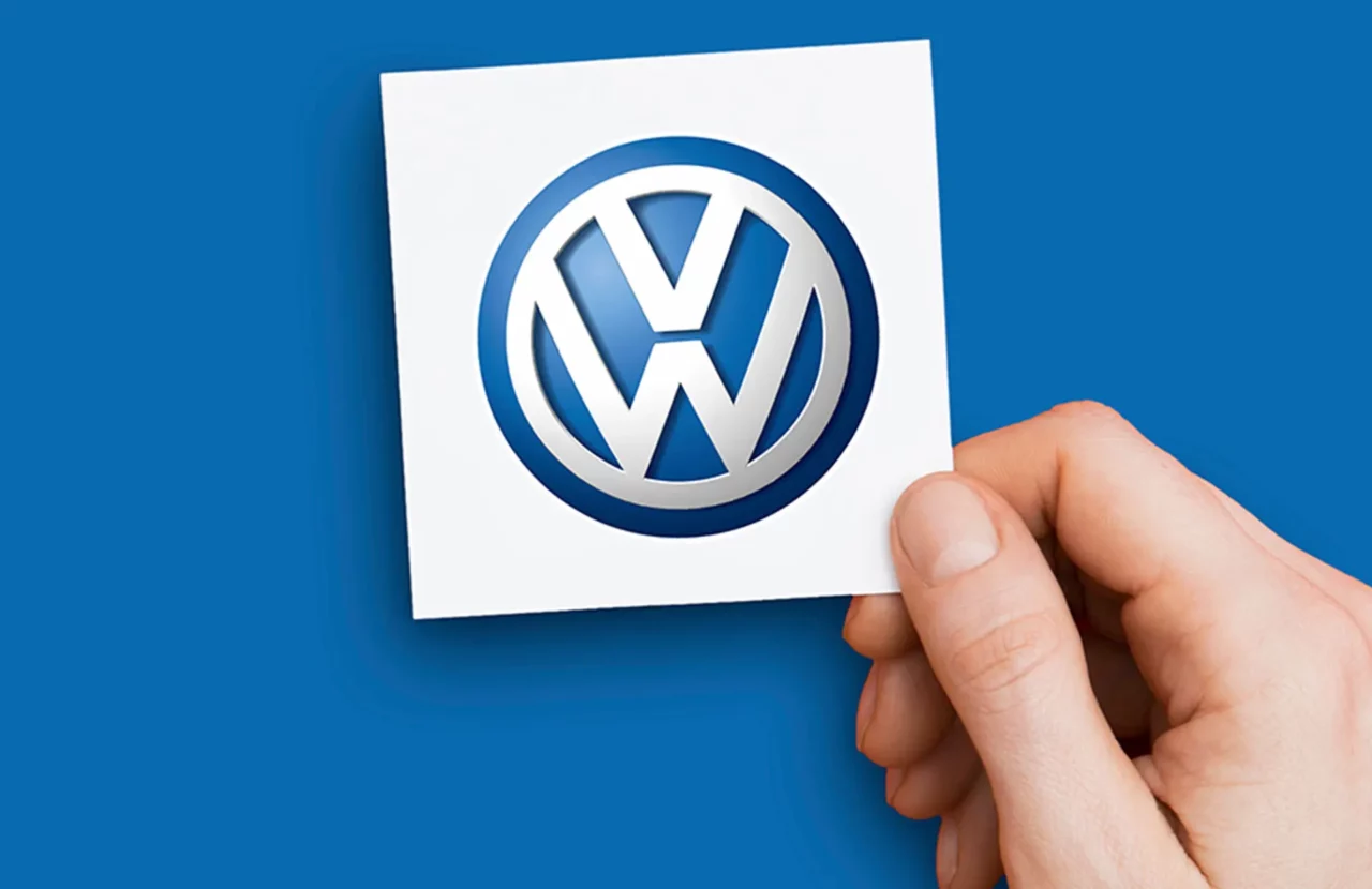 Main tenant une carte blanche avec le logo Volkswagen officiel sur un fond bleu.