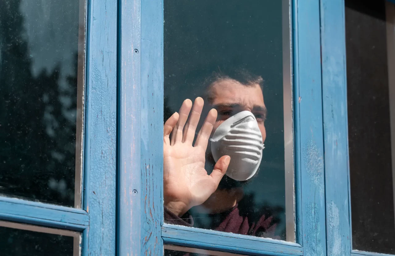 Ein Mann mit medizinischer Maske schaut durch ein Fenster, eine Hand ruht auf der Glasscheibe.