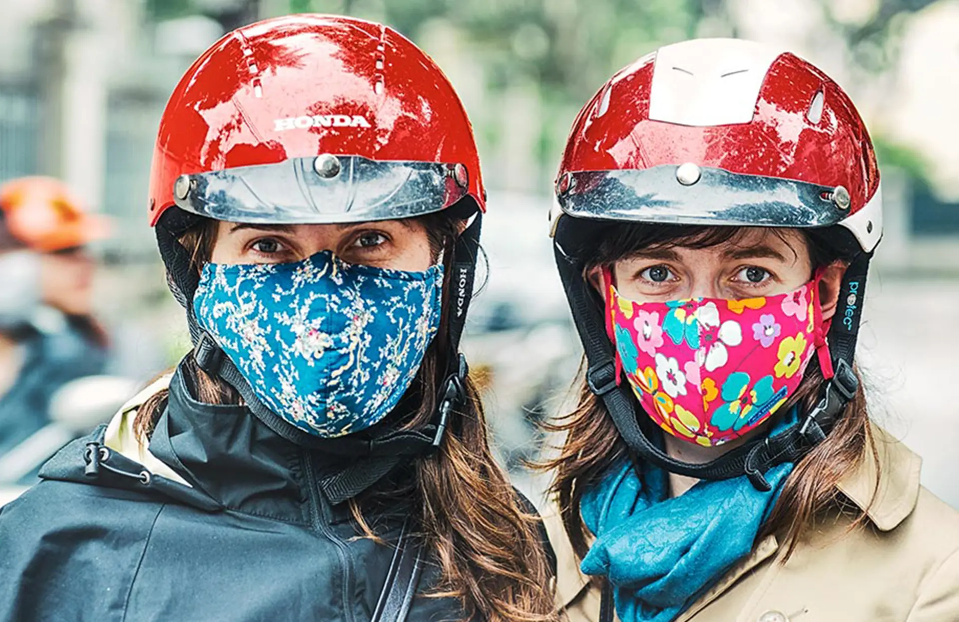 Two women wearing red helmets and colorful masks, ready for a motorcycle ride.