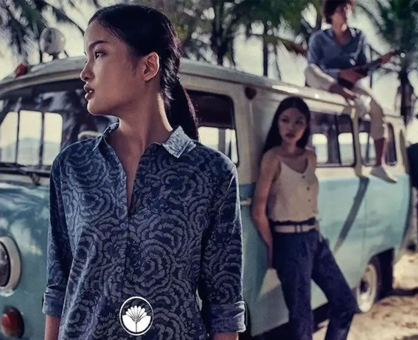 Mujer con una camisa con estampado floral, delante de una furgoneta vintage azul, con otras dos personas al fondo bajo las palmeras.