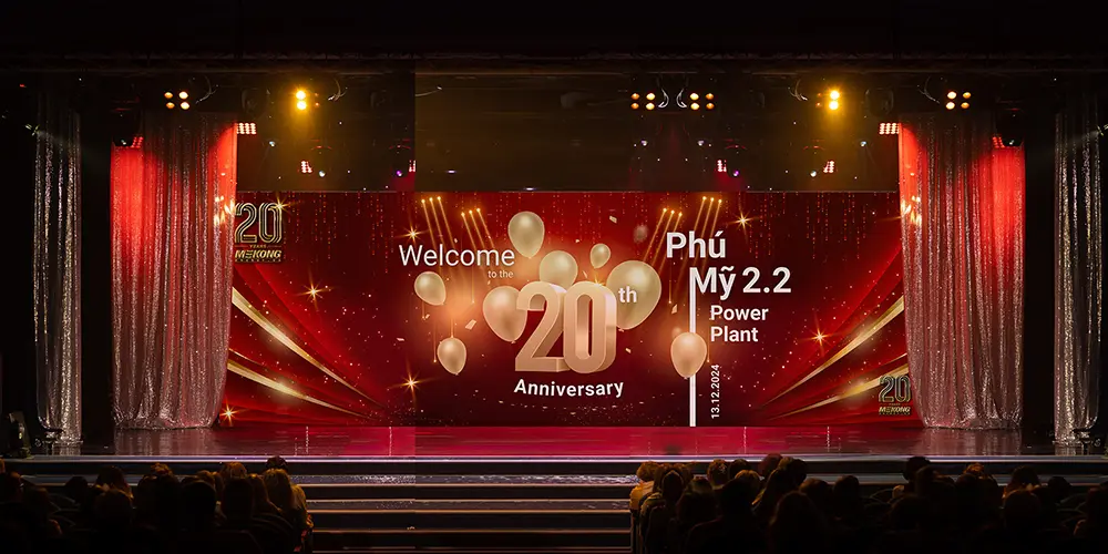 Gran escenario decorado en rojo y dorado con globos dorados y las palabras “Bienvenidos al 20º Aniversario – Planta de Energía Phú Mỹ 2.2” para el evento del 20º Aniversario de Mekong Energy.