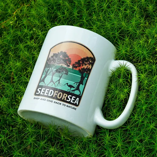 Taza blanca con el logo de Seed for Sea que representa manglares, el mar y una puesta de sol, colocados sobre una alfombra de musgo verde.